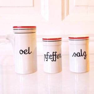 May include: Set of three white ceramic kitchen canisters with black lettering and red accents. The canisters are labeled "oel", "pfeffer", and "salz". The lids have a red band and black trim. The canister on the left has a spout.