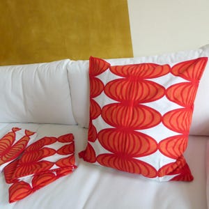 May include: White cushions featuring a repeating pattern of red and orange oval shapes. The cushions are on a white surface, with a mustard-coloured wall in the background. These cushions are a decorative home accent.