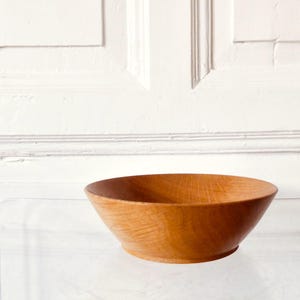Puede incluir: Un cuenco de madera marrón claro con un diseño ancho y poco profundo. El cuenco tiene un acabado liso y pulido y un patrón de vetas de madera natural. El cuenco probablemente se utiliza para servir alimentos o como objeto decorativo.