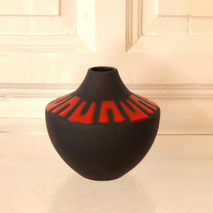 May include: A matte black vase with a rounded body and a narrow neck. The upper shoulder of the vase features a repeating pattern of raised, bright orange U-shaped motifs that encircle the circumference of the piece in a symmetrical design.