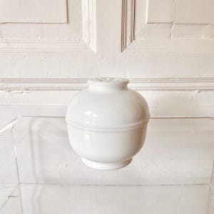 Op de afbeelding: Een kleine, witte keramische vaas met een afgeronde body en een smalle, gegroefde hals. De vaas heeft een gladde, glanzende afwerking en staat op een transparant oppervlak. De achtergrond is een witte muur met panelen.