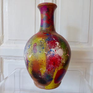 Könnte beinhalten: Eine handbemalte Keramikvase mit bauchigem Körper und schlankem Hals. Die Vase ist mit einem lebendigen abstrakten Design mit Farbspritzern in Rot, Gelb, Grün, Blau und Weiß verziert. Die Vase hat einen braunen Boden und Rand.