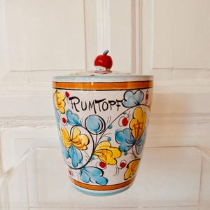 May include: A white ceramic jar with a lid, decorated with yellow and blue floral patterns, and the word "RUMTOPF" in black script. The lid has a red apple-shaped handle. Orange and blue bands encircle the jar.