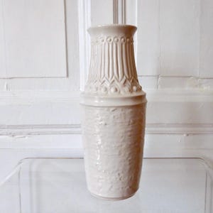Könnte beinhalten: Eine cremefarbene Keramikvase mit strukturierter Oberfläche und dekorativem, geriffeltem Hals. Die Vase hat eine zylindrische Form und steht auf einer transparenten Oberfläche. Es ist ein dekorativer Einrichtungsgegenstand.