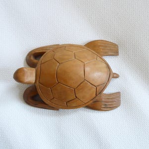 Könnte beinhalten: Eine hellbraune Holzschildkrötenfigur mit detailliertem, segmentiertem Panzer und geschnitzten Flossen und Kopf. Die Schildkröte scheint aus einem einzigen Holzstück gefertigt zu sein. Vor weißem Hintergrund.
