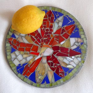 Könnte beinhalten: Ein runder Mosaik-Trittstein mit einem Blumendesign aus roten, orangefarbenen, weißen und blauen Glasstücken. Eine gelbe Zitrone liegt auf dem Stein. Das Mosaik ist von grünen und grauen Fliesen umrandet.