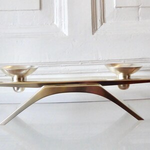 Könnte beinhalten: Ein Kerzenhalter aus Messing im Mid-Century-Modern-Stil mit zwei Kerzenschalen. Der Halter hat ein schlankes, minimalistisches Design mit geschwungenem Fuß und flacher Oberseite. Das Messing hat eine warme, metallische Oberfläche.