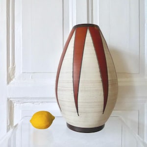 Könnte beinhalten: Eine hohe, cremefarbene Vase mit vertikalen, rotbraunen dreieckigen Akzenten. Die Vase hat einen dunkelbraunen Rand und Boden. Links von der Vase liegt eine gelbe Zitrone. Die Vase hat eine moderne Ästhetik aus der Mitte des Jahrhunderts.