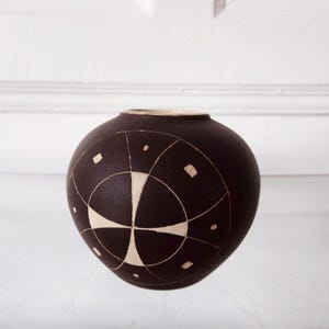 Puede incluir: Un jarrón de cerámica marrón oscuro con forma esférica. Presenta un diseño geométrico en crema con líneas, círculos y cuadrados que se cruzan. El borde del jarrón es de un color más claro.