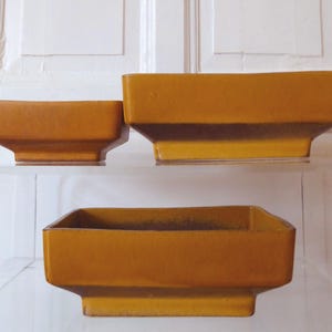 May include: Three rectangular, ceramic planters in varying sizes. The planters are a warm, golden-brown color with a slightly glossy finish. They have a stepped base and a simple, modern design, suitable for indoor or outdoor use.