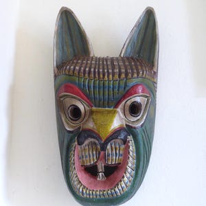 May include: A hand-carved wooden mask featuring a stylised animal face with pointed ears. The mask is painted in shades of green, yellow, red, and black, with a wide-open mouth revealing two prominent front teeth and detailed linear carvings.