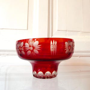 Op de afbeelding: Een helderrode glazen kom met een voet. De kom heeft geëtste bloemmotieven rond de rand en decoratieve sneden op de basis. Een opvallend decoratief stuk.