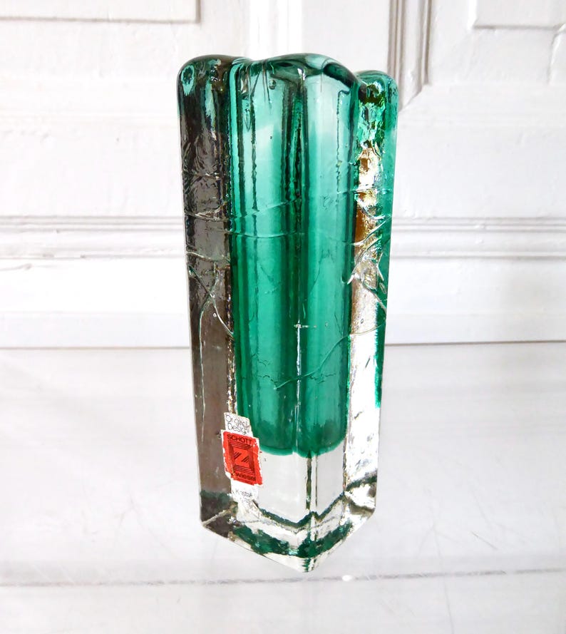 May include: A rectangular, clear glass vase with a teal-green interior. The vase has a textured, cracked appearance on the exterior. A red and white sticker is affixed to the front. The vase is approximately 10 inches tall.