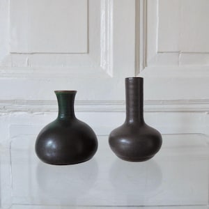 May include: Two dark brown ceramic vases with rounded bodies and narrow necks. One vase has a green rim, the other a brown rim. The vases are displayed on a clear glass surface against a white background.