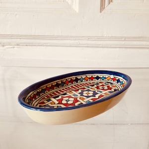 Könnte beinhalten: Eine ovale Keramikschale mit einem handbemalten geometrischen Muster in Blau, Rot, Gelb und Weiß. Das Innere zeigt ein komplexes ineinandergreifendes Stern- und Blumenmotiv, während die Außenseite ein glattes, hellbeiges Finish aufweist.