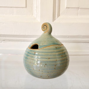 Könnte beinhalten: Handgefertigter Keramiktopf mit hellblauer Glasur und horizontalen Streifen. Der Topf hat einen runden Körper, eine kleine Öffnung und einen spiralförmigen Griff oben. Der Topf ist etwa 15 cm hoch.