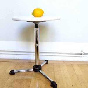 Pode incluir: Uma pequena mesa de altura ajustável, com um tampo branco e uma base de metal cromado com três pernas. Um limão amarelo está sobre a superfície da mesa. A altura da mesa é ajustável.