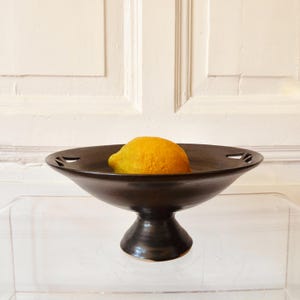 Könnte beinhalten: Eine dunkelbraune Obstschale aus Keramik mit Sockel, die eine leuchtend gelbe Zitrone enthält. Die Schale hat dekorative Aussparungen am Rand. Die Schale steht auf einer transparenten Oberfläche.