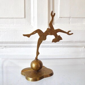 Könnte beinhalten: Eine Messingfigur einer Person in dynamischer Pose, die auf einem Fuß auf einer kugelförmigen Basis balanciert. Die Figur weist geätzte Liniendetails am Körper und an der Kleidung auf und ist auf einer flachen, unregelmäßig geformten Basis montiert.