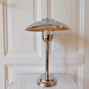 May include: A chrome-finished table lamp with a domed shade and a slender, vertical stem. The lamp features a circular base and a small, light green accent at the top. The interior of the shade has a gold finish, and the lamp is on a clear surface.