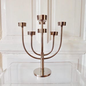 Könnte beinhalten: Ein silberfarbener Kerzenleuchter mit sieben Kerzenhaltern. Der Kerzenleuchter hat einen kreisförmigen Sockel und einen zentralen Stiel mit gebogenen Armen zur Aufnahme der Kerzen. Das Design ist symmetrisch und elegant, geeignet für die Inneneinrichtung.