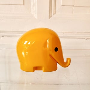Könnte beinhalten: Eine hellgelbe, stilisierte Elefantenfigur mit rundem Körper und gebogenem Rüssel. Der Elefant hat ein kleines schwarzes Auge und besteht aus glänzendem Material. Die Figur ist ein dekoratives Objekt.