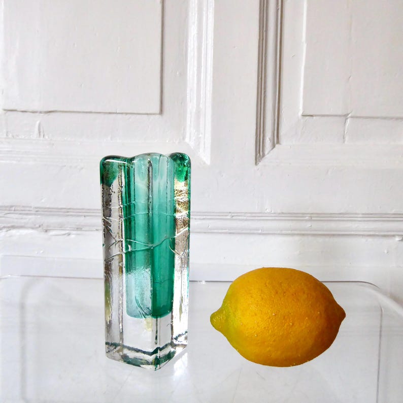 May include: A rectangular, clear glass vase with a teal interior and a textured exterior. A yellow lemon sits to the right of the vase. The vase has a unique, artistic design, perfect for displaying flowers or as a decorative piece.