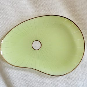 Può includere: Un vassoio da portata asimmetrico a forma di rene di colore verde lime chiaro, caratterizzato da un foro circolare centrale e da un delicato bordo color oro. La superficie è decorata con sottili linee radianti che partono dal centro.