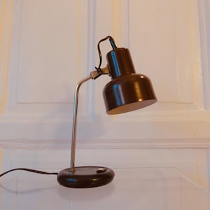 Puede incluir: Una lámpara de escritorio marrón con un brazo curvo plateado y una base redonda. La lámpara tiene una pantalla en forma de campana marrón oscuro y un cable de alimentación negro. La lámpara está sobre una superficie transparente.
