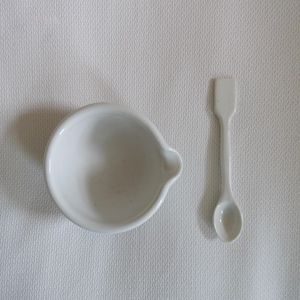 Könnte beinhalten: Ein weißes Keramik-Mörser- und Stößel-Set mit einem kleinen Ausguss am Mörser. Ein weißer Keramikspatel befindet sich neben dem Mörser. Das Set befindet sich auf einer weißen strukturierten Oberfläche.