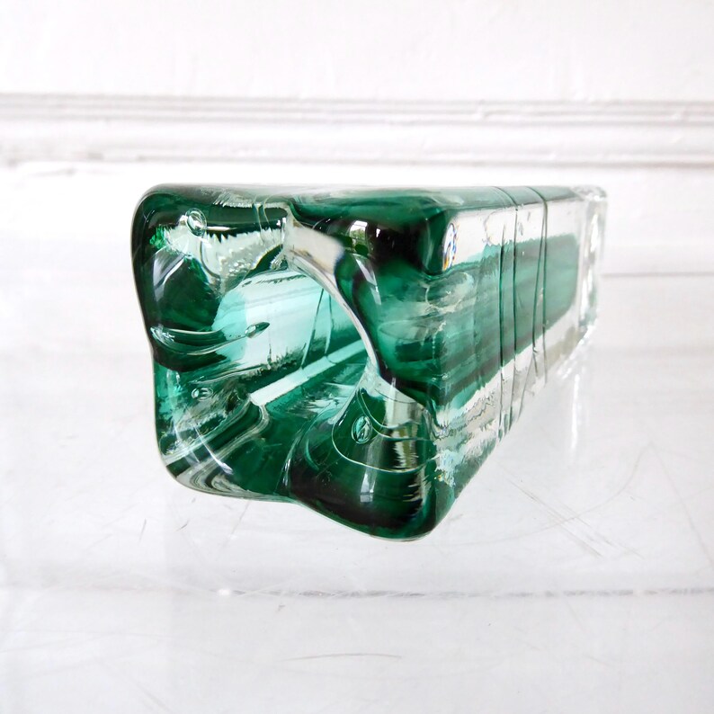 May include: A rectangular, emerald green glass vase with a unique, curved opening. The vase has a transparent quality, allowing light to pass through, and is suitable for displaying flowers or as a decorative piece. The vase is approximately 10 inches long.