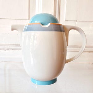 Puede incluir: Una tetera de cerámica blanca con cuerpo redondeado, asa curva y pico corto. La tapa es una cúpula lisa de color azul claro. Una banda horizontal en azul claro y una fina franja naranja rodean la parte superior del cuerpo de la tetera.