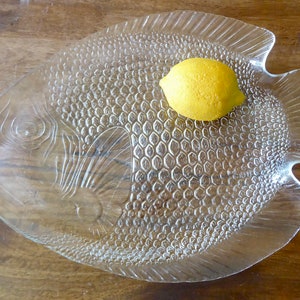 Puede incluir: Un plato de servir de vidrio transparente con forma de pez y superficie texturizada. Un limón amarillo se encuentra sobre el plato.