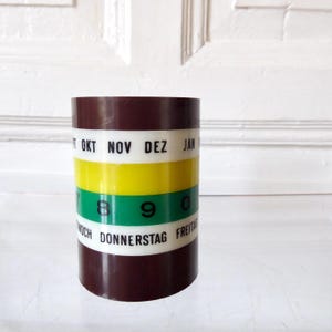 Può includere: Un calendario cilindrico vintage con parte superiore e inferiore marrone scuro. Il calendario presenta fasce orizzontali bianche, gialle e verdi, con i mesi e i giorni della settimana stampati sulle fasce. I numeri 7, 8, 9 e 0 sono visibili.