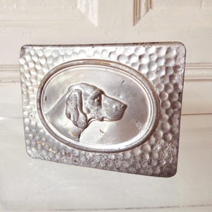May include: A rectangular, silver-colored metal plaque with a hammered texture. An oval in the center features a detailed profile of a dog's head. The dog's features are clearly defined, and the metal has a slightly aged appearance.
