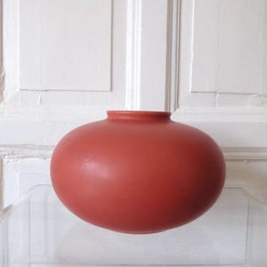 Könnte beinhalten: Eine terracottafarbene, runde Vase mit schmalem Hals. Die Vase hat eine glatte, matte Oberfläche und ein schlichtes Design. Der Hintergrund ist eine weiße, getäfelte Oberfläche.