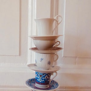 May include: A stack of four teacups and saucers. The bottom cup is decorated with a colorful floral pattern. The second cup is white with blue flowers. The third cup is white with a gold rim. The top cup is white with a gold handle.
