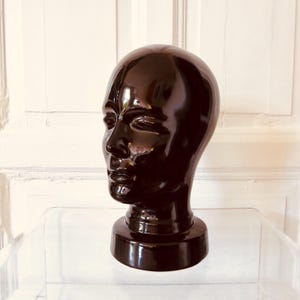 Könnte beinhalten: Eine glänzende, dunkelbraune Keramikkopfskulptur. Die Skulptur zeigt ein detailliertes Gesicht mit geschlossenen Augen, einer definierten Nase und vollen Lippen. Der Kopf sitzt auf einem zylindrischen Sockel.