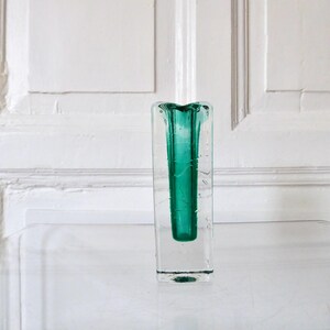 May include: A rectangular clear glass vase with a vibrant teal interior. The vase has a square base and a slightly flared top. The background is a white wall with paneling. The vase is sitting on a clear surface.