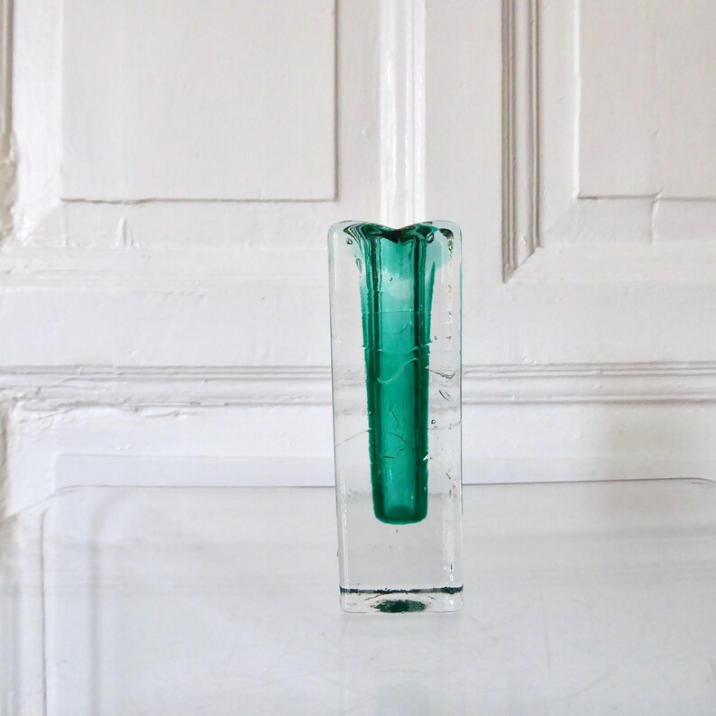 May include: A rectangular clear glass vase with a vibrant teal interior. The vase has a square base and a slightly flared top. The background is a white wall with paneling. The vase is sitting on a clear surface.