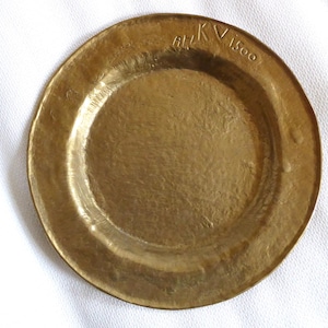 May include: A round, antique brass plate with a hammered texture. The plate has a slightly raised rim and a shallow center. The text "Ah/KV 1500" is stamped on the rim. The plate is a warm, golden color.
