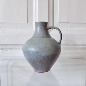 May include: A light grey ceramic vase with a textured surface and a handle. The vase has a rounded body and a narrow neck.