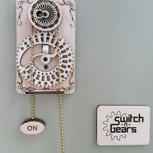 May include: A wooden light switch with a gear mechanism and two pull chains labeled "ON" and "OFF". The switch is mounted on a light beige wall with a small wooden sign that reads "Switch-n-Gears".