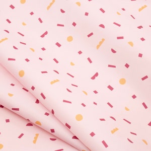 May include: A light pink fabric with a repeating pattern of geometric shapes in shades of burgundy and yellow. The shapes include rectangles, circles, and semi-circles, creating a playful and modern design. The fabric is folded, showing its texture and drape.