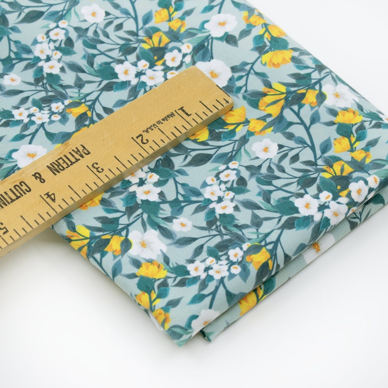 May include: A close-up of a light blue fabric with a white and yellow floral pattern. The fabric is folded and a ruler is placed on top of it. The ruler is marked with inches and the text "Made in USA".