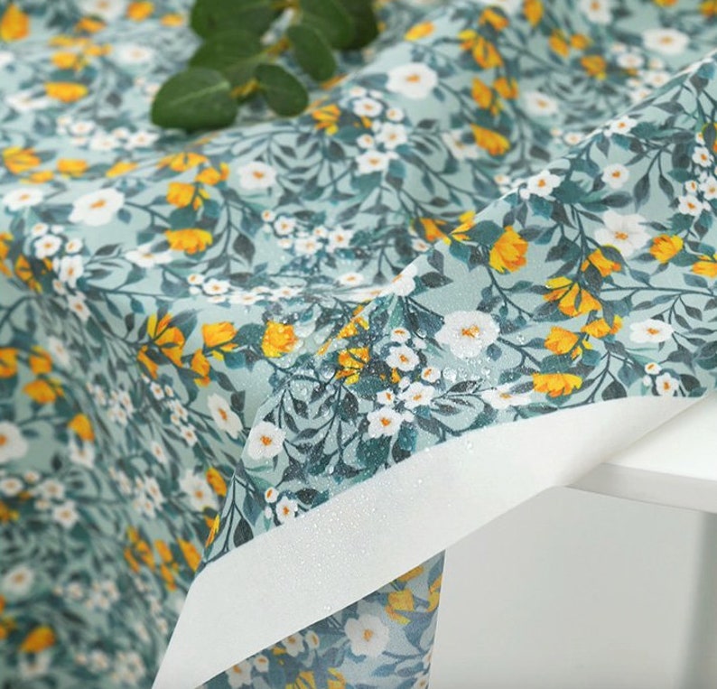 May include: A close-up of a light blue fabric with a white and yellow floral pattern. The fabric is waterproof and has a subtle sheen.