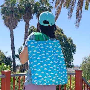 May include: A person wearing a light blue hat with a green strap is carrying a blue tote bag with a white cloud pattern. The bag is made of a lightweight, foldable material.