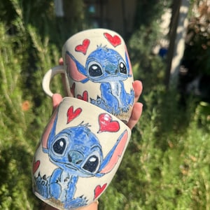 May include: Two ceramic mugs with a blue cartoon character design, pink ears, and red hearts. The mugs are hand-painted with a whimsical design, ideal for tea or coffee. The mugs are a unique addition to any kitchen.
