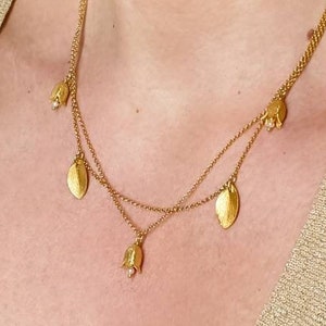 Könnte beinhalten: Eine goldene mehrreihige Halskette mit Blatt- und Blumenanhängern. Die Kette wird von einer Person mit einem hellbraunen Pullover getragen.