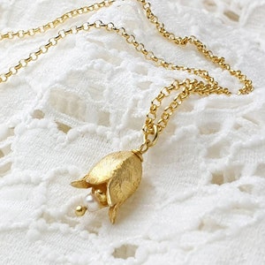 Könnte beinhalten: Eine zarte goldene Halskette mit einem kleinen, strukturierten Blumenanhänger und einem einzelnen Perlenakzent. Die Halskette liegt auf einem weißen Spitzenhintergrund.